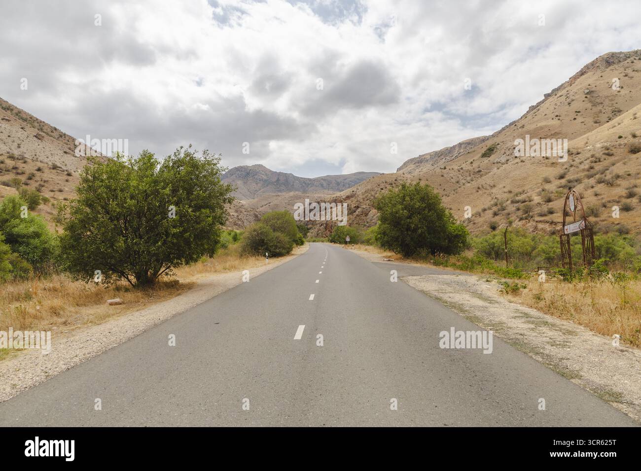 Vue d'une route asphaltée serpentant à travers des montagnes arides sous un ciel nuageux, encadrée par des arbres clairsemés et une végétation sèche, monastère de Noravank, province de Vayots Dzor, Arménie. Banque D'Images