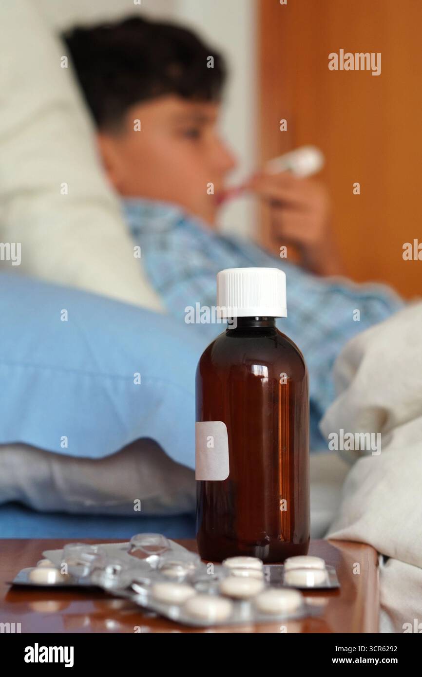 enfant en pyjama malade au lit avec le thermomètre en place et les médicaments sur la table de chevet Banque D'Images