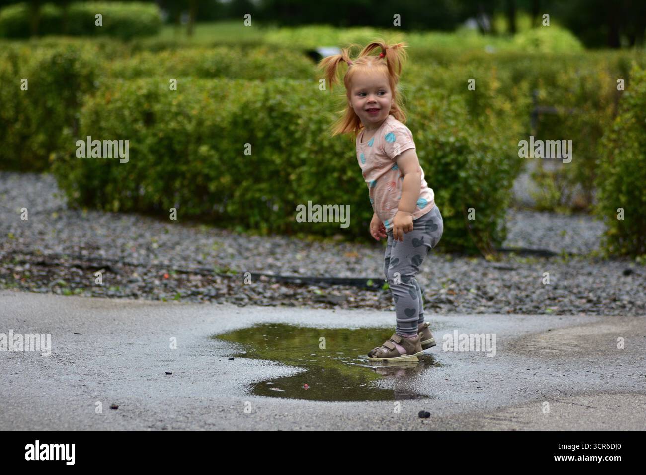 Mignon jeune fille aux cheveux rouges avec des queues de cochon souriant tout en se tenant dans une flaque sur un trottoir mouillé. Enfant jouissant de jeux en plein air, amusant éclaboussure d'eau et happ Banque D'Images