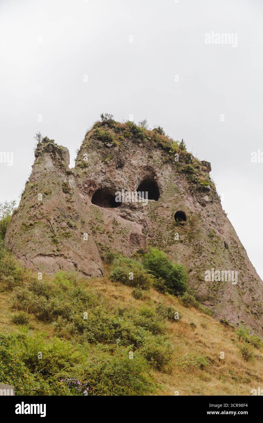 Vue d'une montagne rocheuse altérée surmontée d'une végétation clairsemée et sculptée avec des ouvertures sombres de grotte, sous un ciel sourd, Goris, province de Syunik, Arménie. Banque D'Images