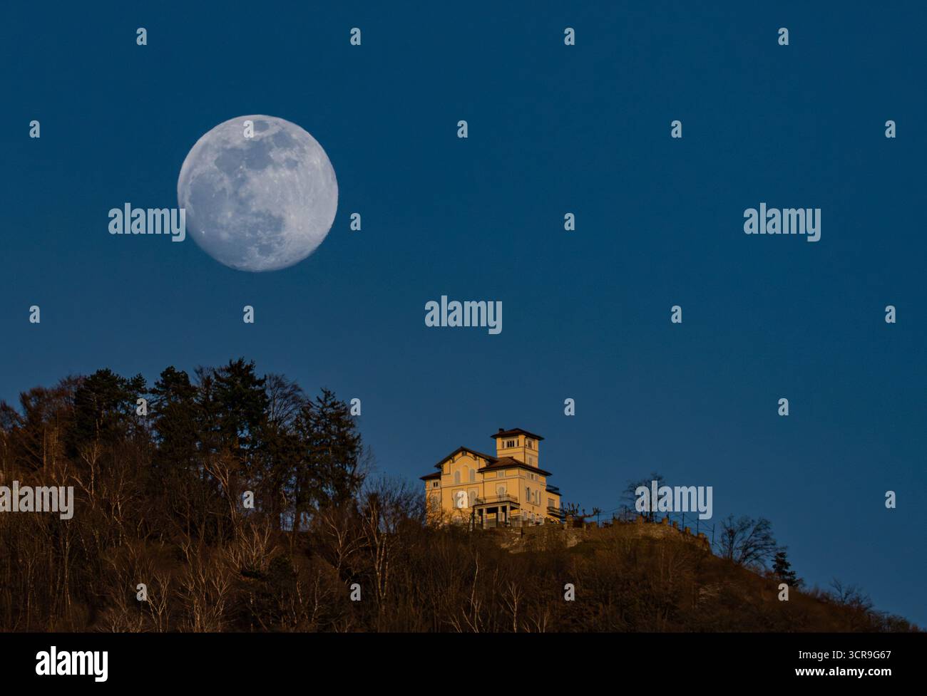 Vue aérienne d'une pleine lune lumineuse accroche haut au-dessus d'un bâtiment doré perché au sommet d'une colline, sa silhouette frappée contre le ciel assombrissant, Lugano, Suisse Banque D'Images