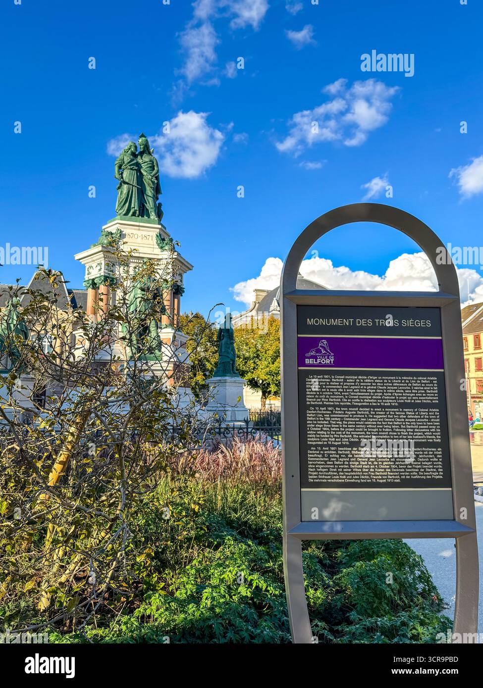 Vue sur le Monument des trois sièges avec un panneau d'information au premier plan, situé dans le centre historique de Belfort sous un ciel bleu clair - Image de stock capturée avec un smartphone