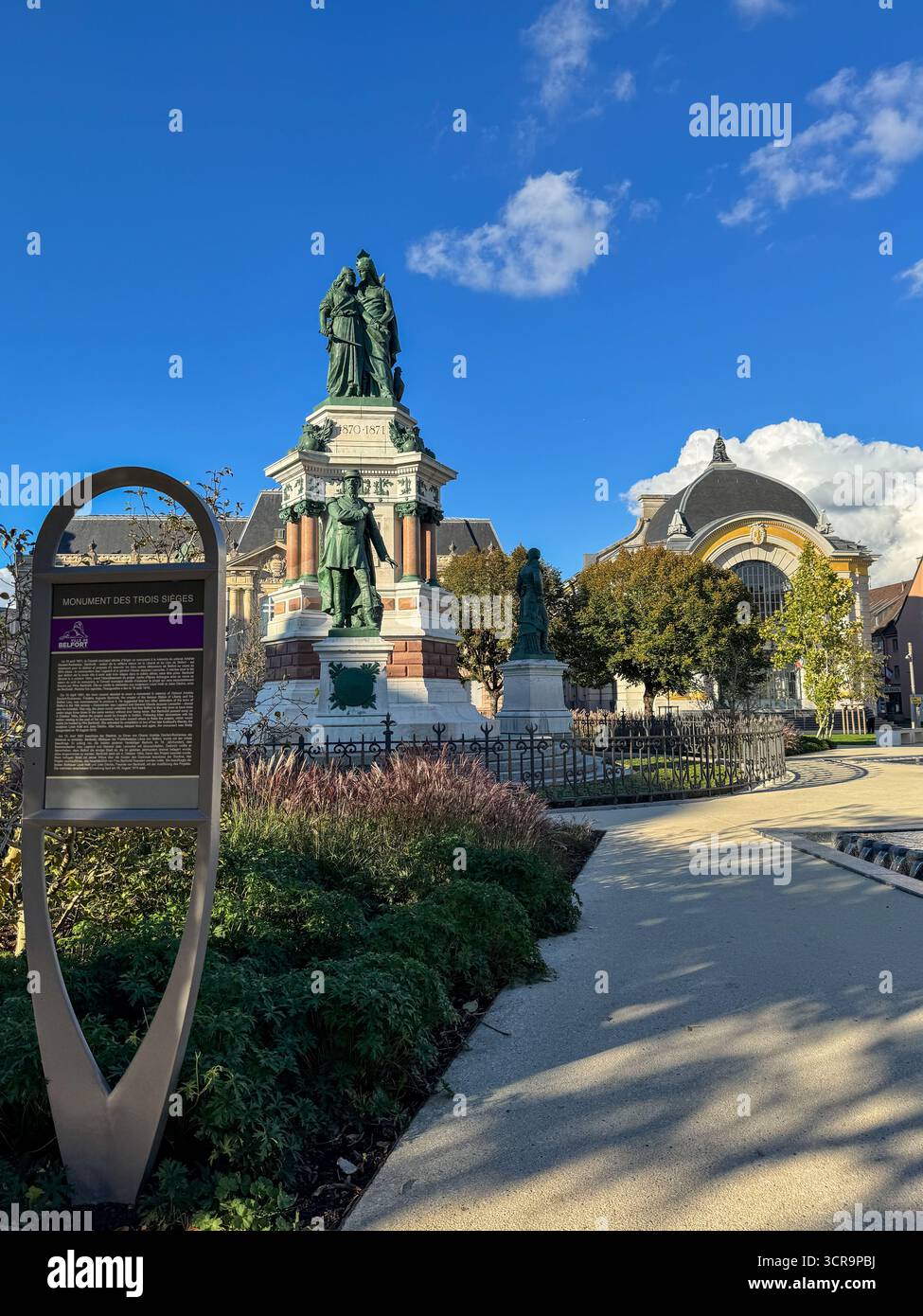 Vue sur le Monument des trois sièges avec un panneau d'information au premier plan, situé dans le centre historique de Belfort sous un ciel bleu clair - Image de stock capturée avec un smartphone