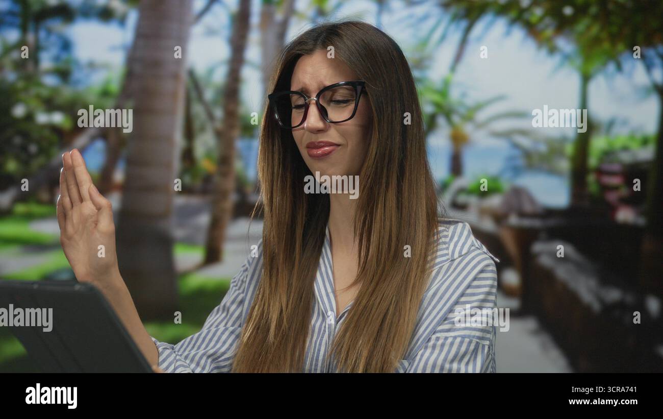 Femme tenant la tablette avec la main levée dans le parc vert parmi les palmiers portant une chemise rayée ; rejet dégoût aversion. Banque D'Images