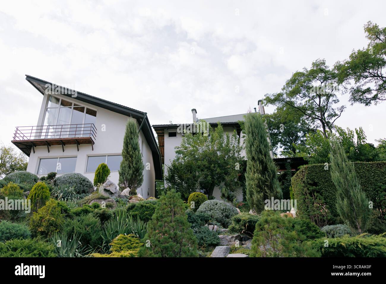 Maison blanche contemporaine avec de grandes fenêtres et un balcon, entourée d'un magnifique paysage en couches avec des conifères, des arbustes et des pierres Banque D'Images