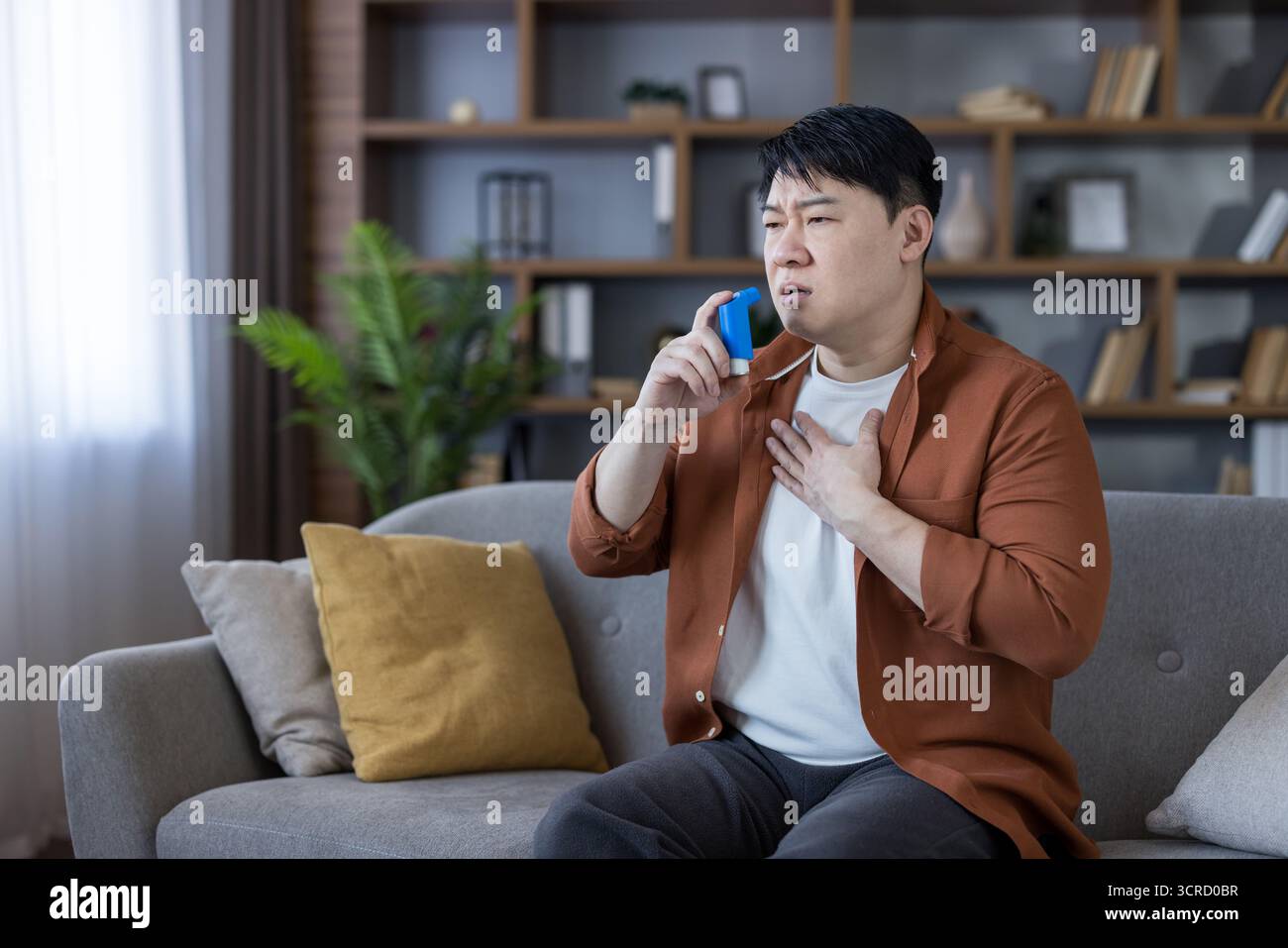 Homme asiatique assis sur un canapé à la maison à l'aide d'un inhalateur bleu, éprouvant un essoufflement et des difficultés à respirer pendant une crise d'asthme, tenant une main sur sa poitrine Banque D'Images