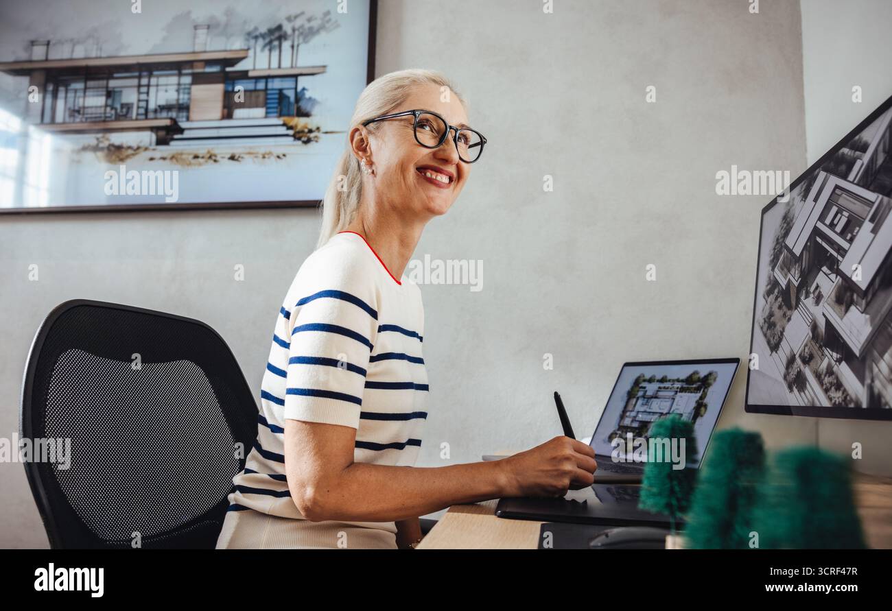 Une architecte senior sourit tout en travaillant à son bureau dans un bureau contemporain avec des outils modernes et des modèles architecturaux. L'image capture pro Banque D'Images