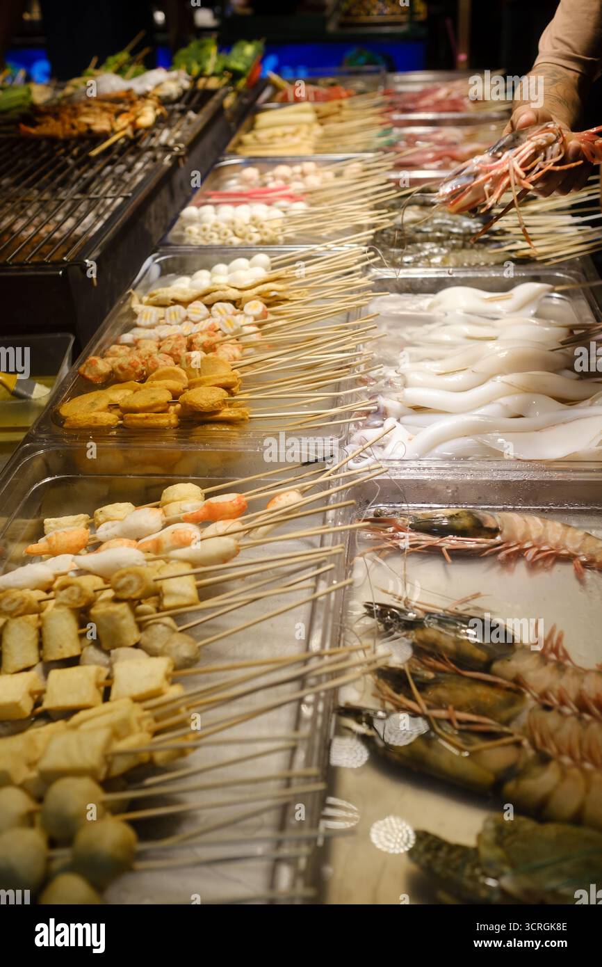 Un étal de marché est rempli d'un assortiment de brochettes de fruits de mer et de viandes. Un vendeur prépare des brochettes fraîches dans une atmosphère animée pendant un même Banque D'Images