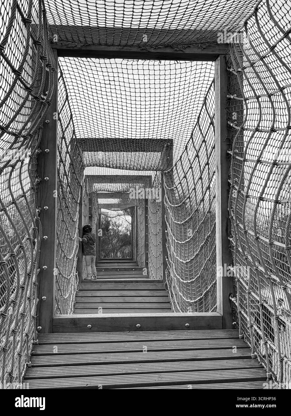 Photo en noir et blanc montrant un enfant sur un pont en bois entouré d'un filet, créant un effet répétitif de tunnel à travers de multiples ouvertures encadrées, Banque D'Images