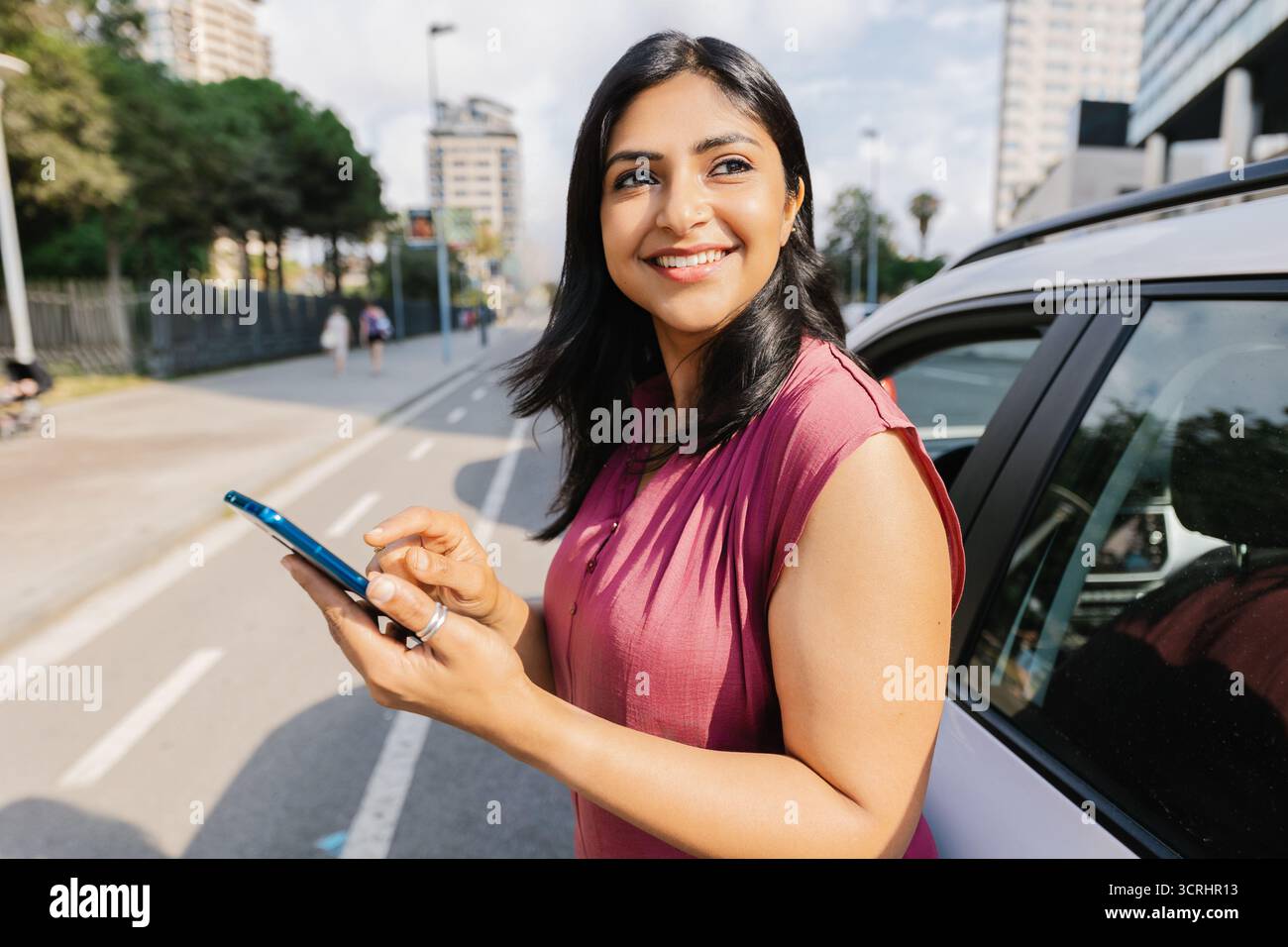Femme indienne souriante à l'aide d'un smartphone à côté de la voiture Banque D'Images Femme indienne souriante à l'aide d'un smartphone à côté de la voiture Banque D'Images
