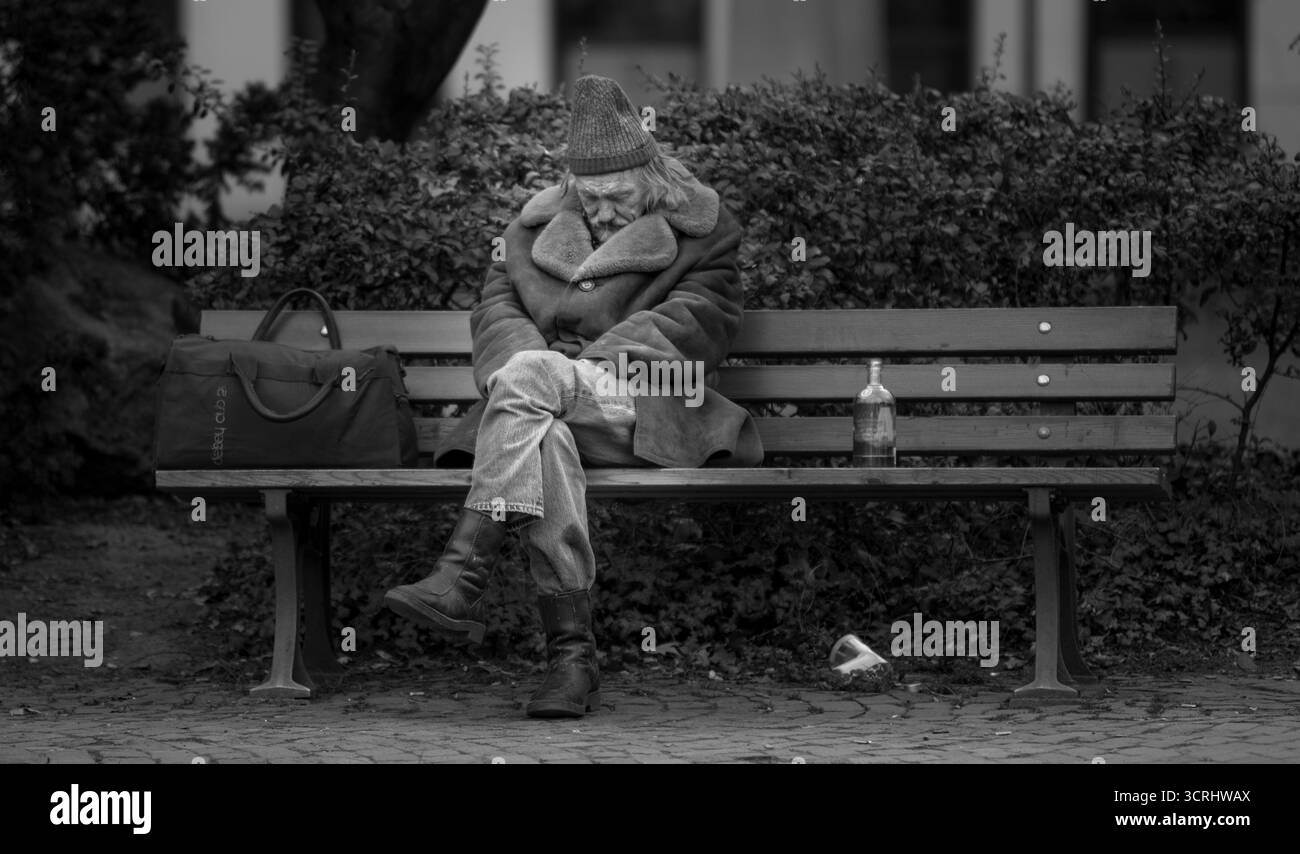 Homme sans-abri dormant sur le banc du parc en manteau d'hiver, photographie de rue en noir et blanc reflétant la solitude et la pauvreté Banque D'Images