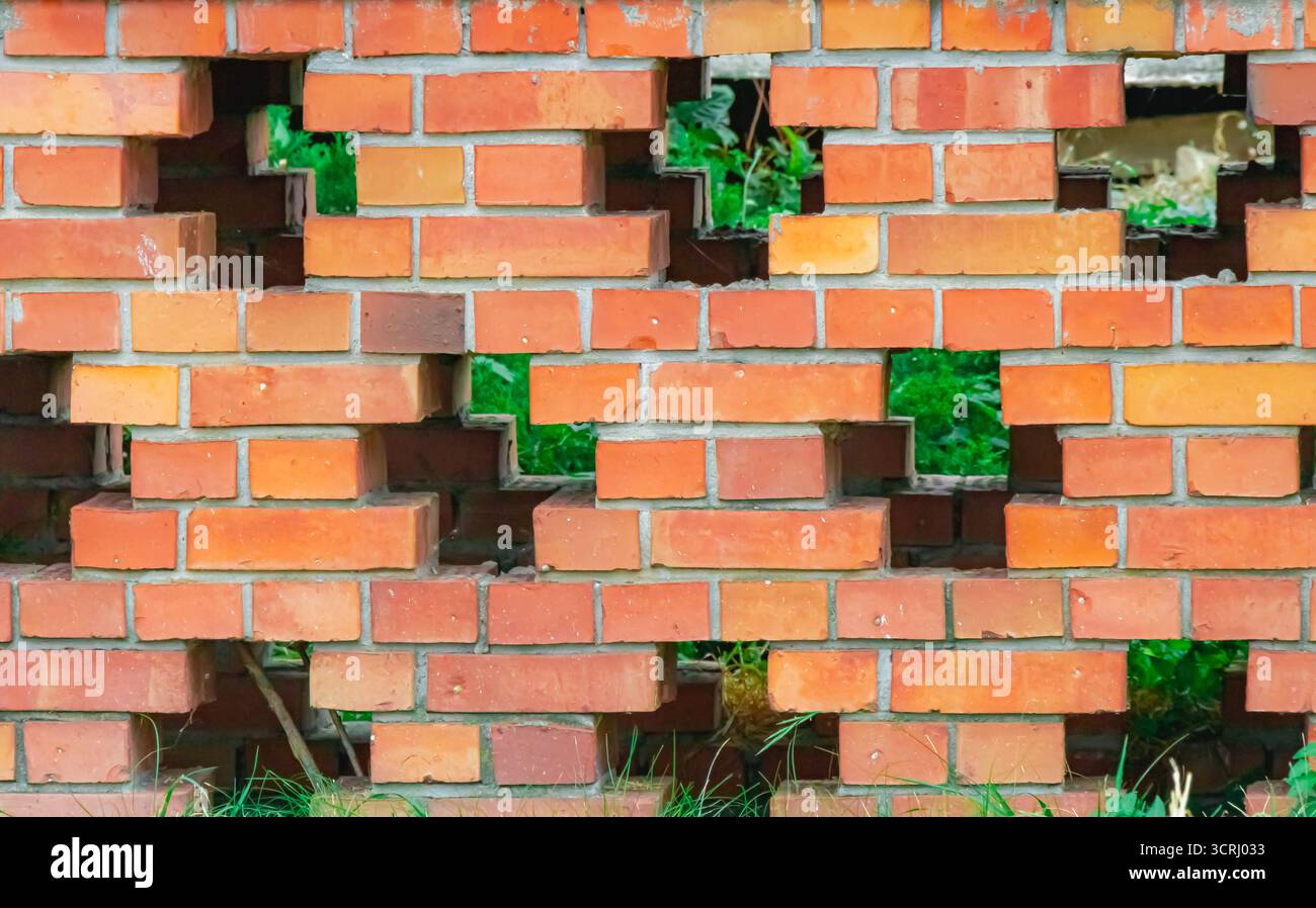 Un mur de briques rouges avec des espaces régulièrement espacés montre des taches d'herbe verte et des plantes derrière, créant un contraste géométrique entre l'architecture et le nat Banque D'Images