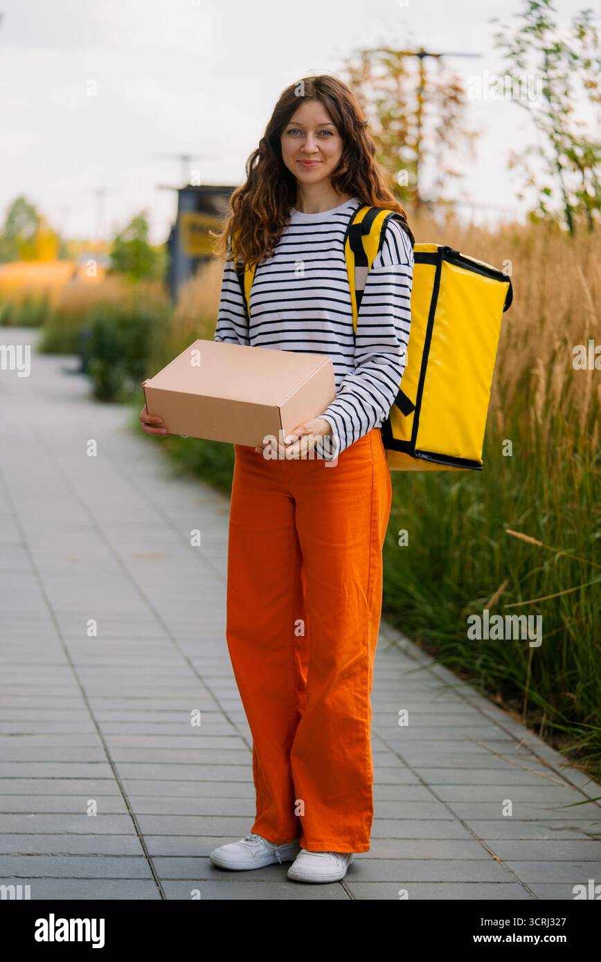 Jeune femme souriante travaillant comme coursier, portant un pantalon orange vif et une chemise rayée, portant un paquet de carton vierge et un grand sac thermique jaune, livrant de la nourriture ou des colis Banque D'Images