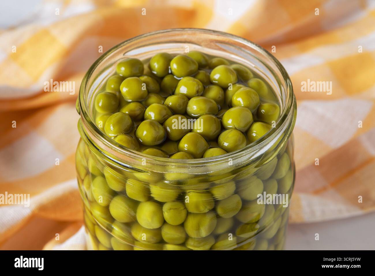 Ouvrez un pot de pois verts en conserve en gros plan. Pot en verre plein de petits pois biologiques marinés sur serviette de cuisine à carreaux. Ingrédient de salade, légume. Banque D'Images