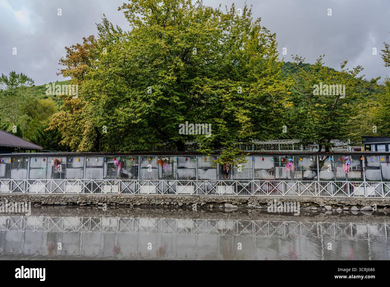 Une clôture décorée au bord de la rivière avec des ornements suspendus colorés et de grands arbres verts reflétés dans l'eau à Qabala, Azerbaïdjan. Banque D'Images