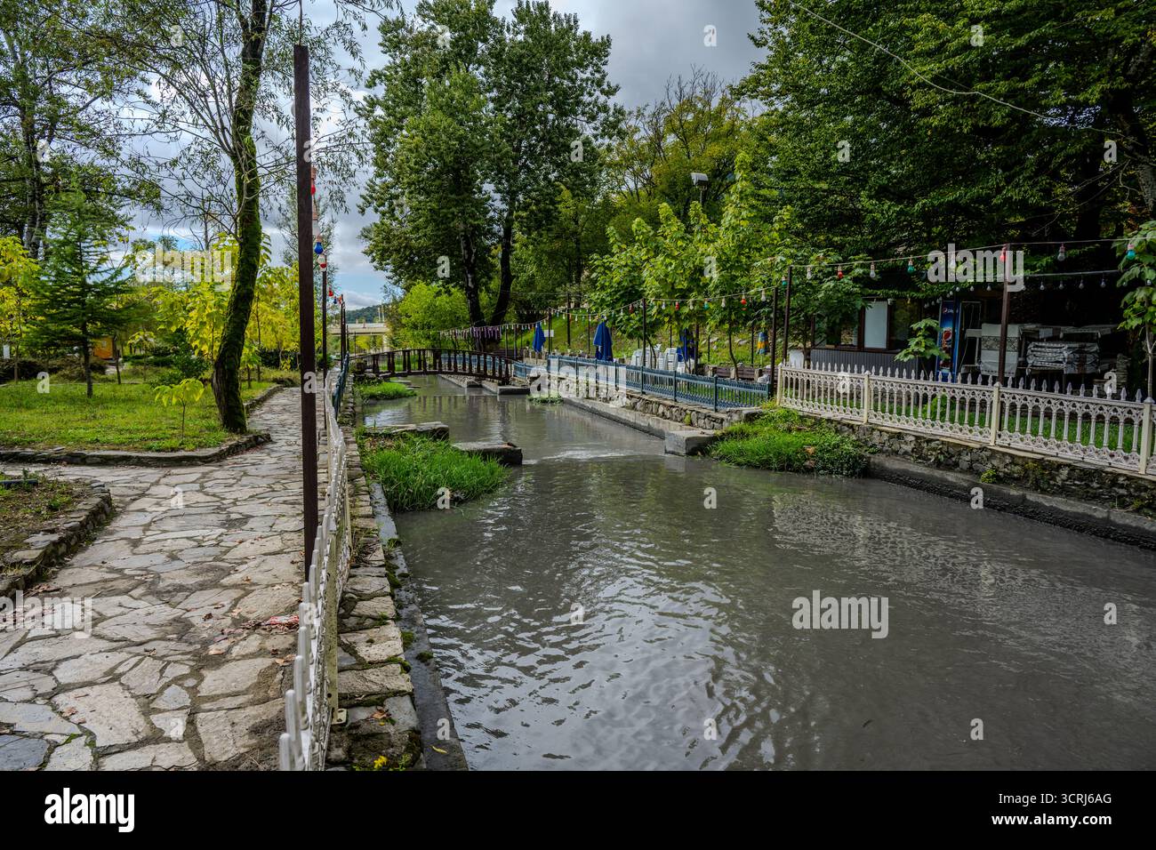 Une passerelle en pierre longe un ruisseau étroit avec des clôtures, des arbres et des places assises en plein air dans un parc de Qabala, en Azerbaïdjan. Banque D'Images