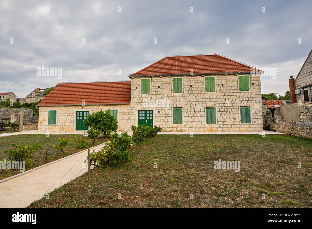 Une maison en pierre de deux étages avec des volets en bois vert se trouve dans une cour rurale tranquille. Toit de tuiles rouges, chemin de gravier et petits arbustes créent un calme et pittoresque Banque D'Images