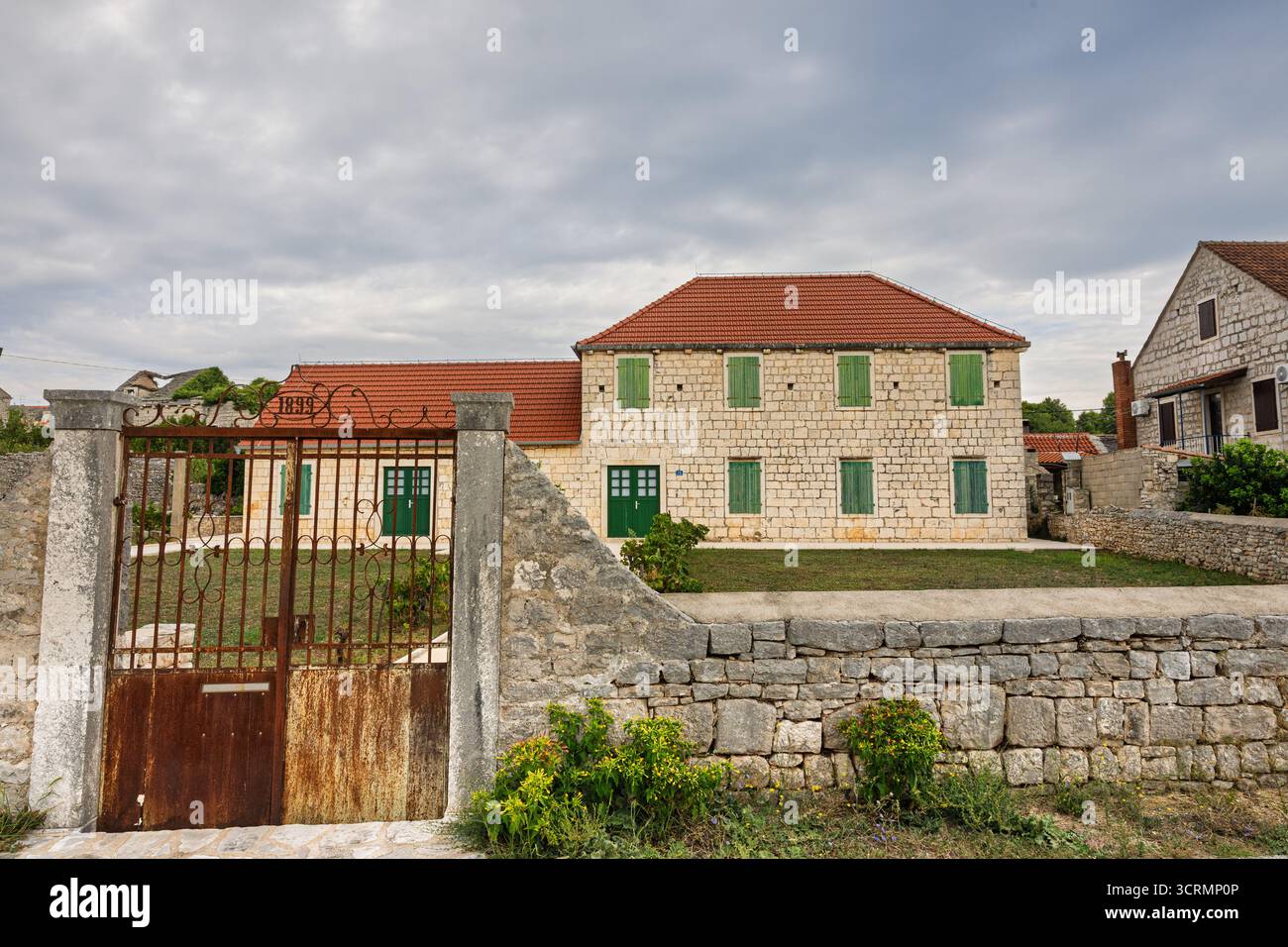 Une maison en pierre de deux étages avec des volets en bois vert se trouve dans une cour rurale tranquille. Toit de tuiles rouges, chemin de gravier et petits arbustes créent un calme et pittoresque Banque D'Images