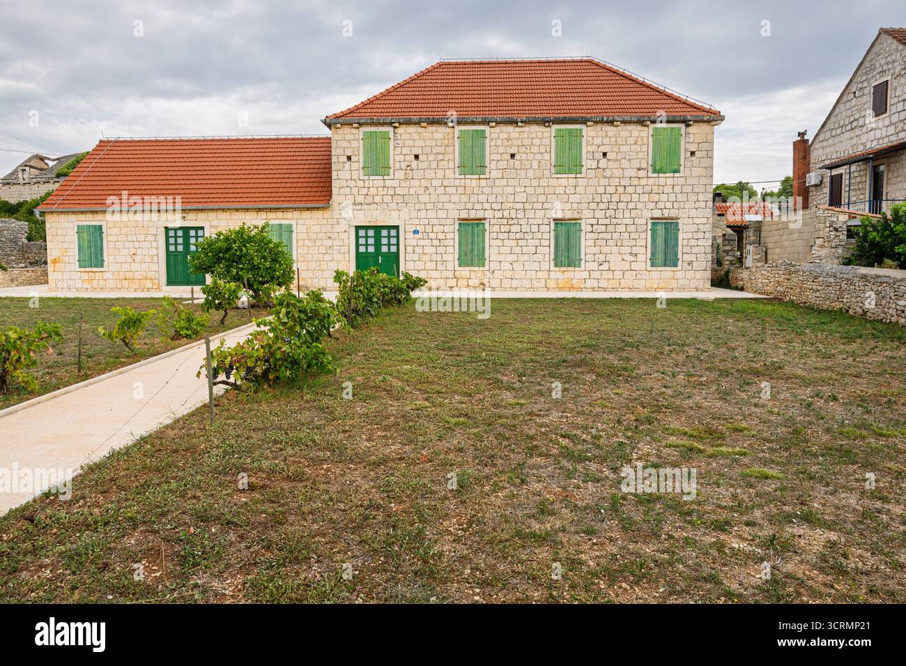 Une maison en pierre de deux étages avec des volets en bois vert se trouve dans une cour rurale tranquille. Toit de tuiles rouges, chemin de gravier et petits arbustes créent un calme et pittoresque Banque D'Images