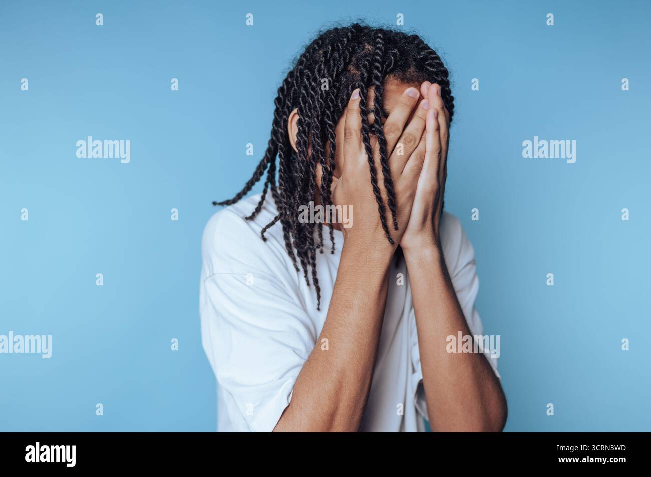 Un jeune homme aux longs cheveux tordus couvre son visage des deux mains. Il est dans un cadre intérieur calme avec un fond bleu doux, véhiculant des émotions profondes. Banque D'Images