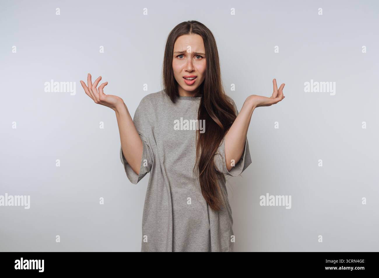 Une jeune femme aux cheveux longs dans une chemise grise se tient sur un fond Uni, exprimant la confusion ou l'incrédulité avec ses bras levés et paumes levées. Banque D'Images