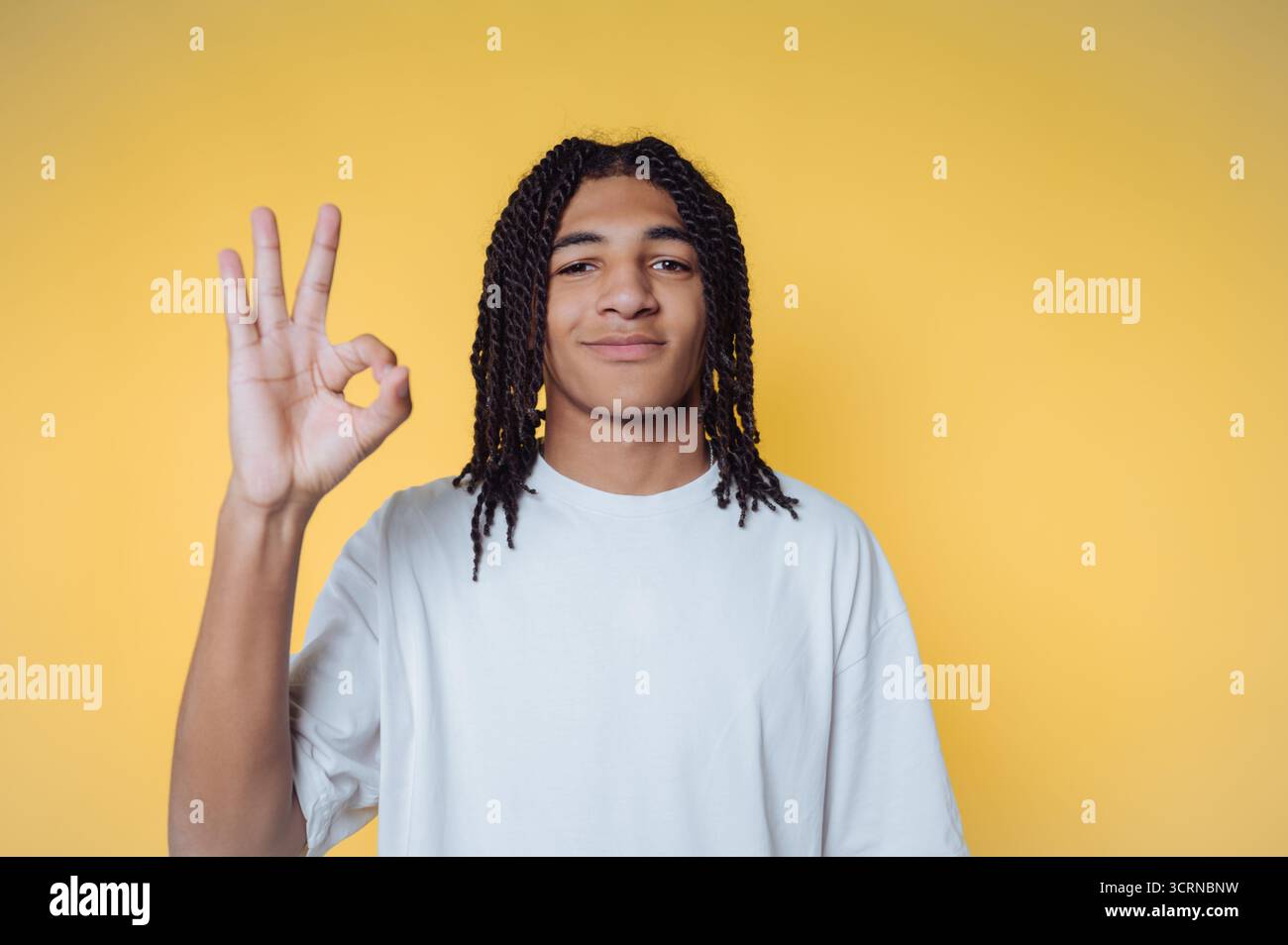 Portrait de jeune homme avec des tresses montre un signe OK, rayonnant positivité sur fond jaune Banque D'Images
