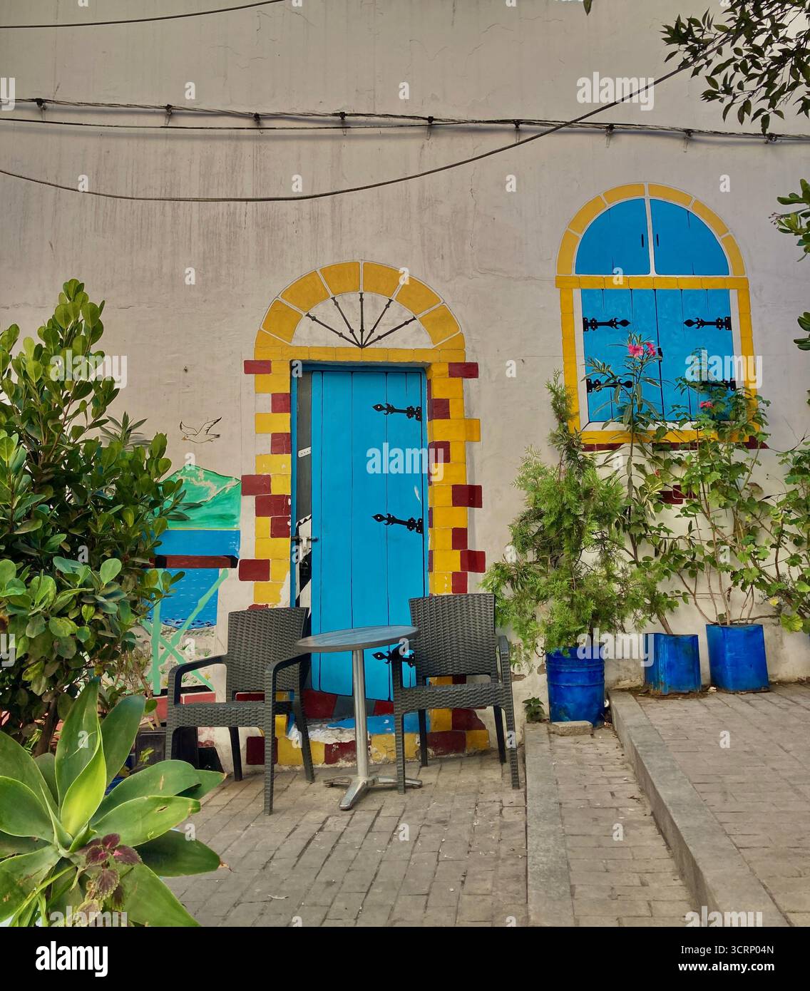 Mur peint avec porte et fenêtre bleues, vraies chaises et plantes à Tanger Kasbah Medina, Maroc. Mélange artistique capturé le 1er octobre 2025. Banque D'Images