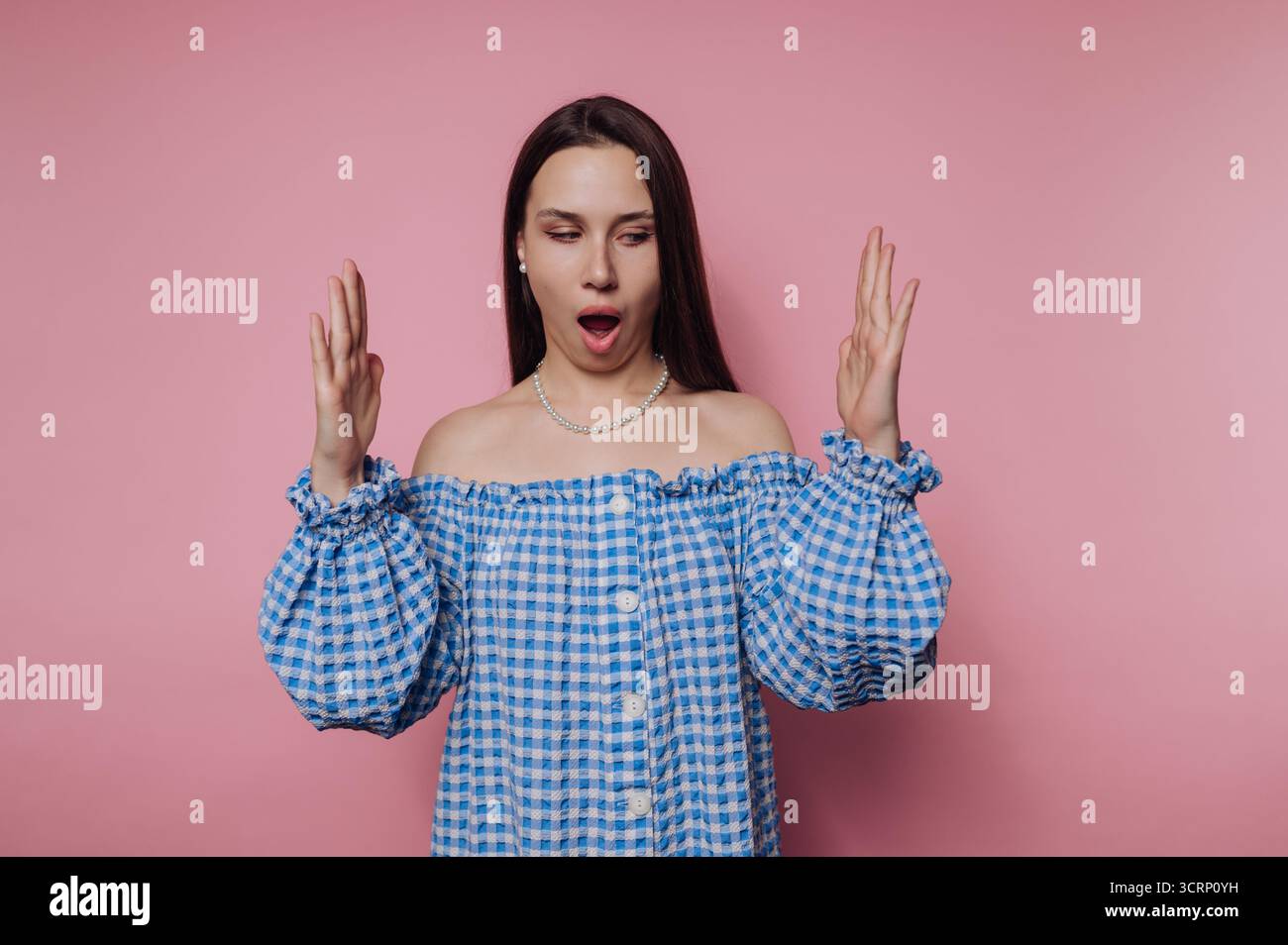Une femme aux longs cheveux foncés porte une blouse bleue à carreaux et montre une expression de surprise. Elle se tient devant un fond rose Uni, mains R Banque D'Images