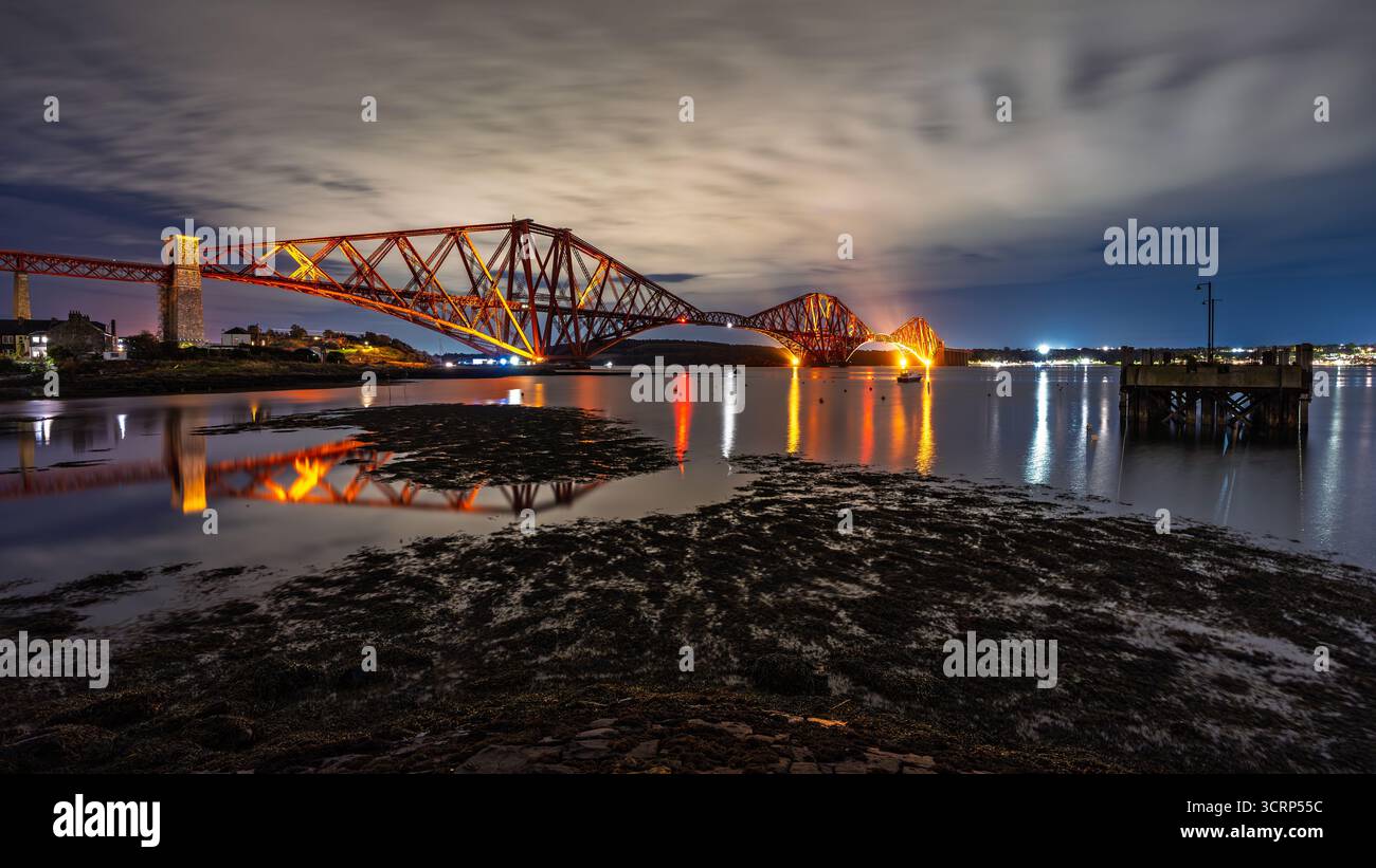 Forth Bridge illuminé, projetant un reflet vibrant sur l'eau avec des nuages en mouvement créant un léger flou de mouvement à marée basse Banque D'Images