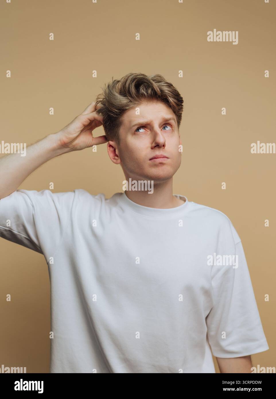 Un jeune homme portant un t-shirt blanc Uni apparaît profondément dans la pensée. Il se gratte la tête, regardant vers le haut avec une expression de contemplation, set aga Banque D'Images