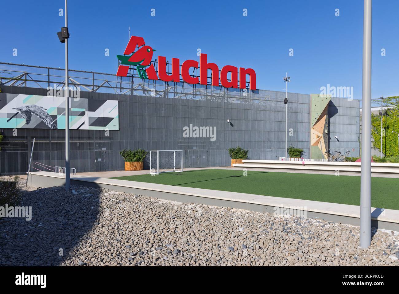 Le logo de l'hypermarché Auchan est bien visible sur l'extérieur sombre et moderne d'un grand magasin. Le premier plan présente un petit soccer synthétique p Banque D'Images