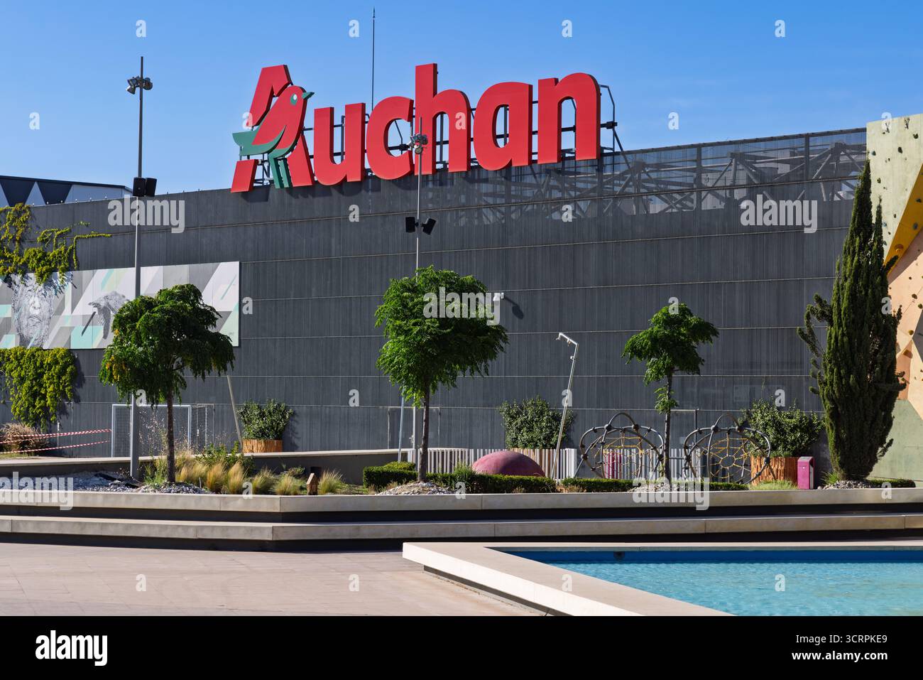 Le grand logo Auchan rouge se trouve au sommet d'un bâtiment commercial moderne et gris. Le premier plan présente des arbres décoratifs, des jardinières et une petite pièce d'eau Banque D'Images
