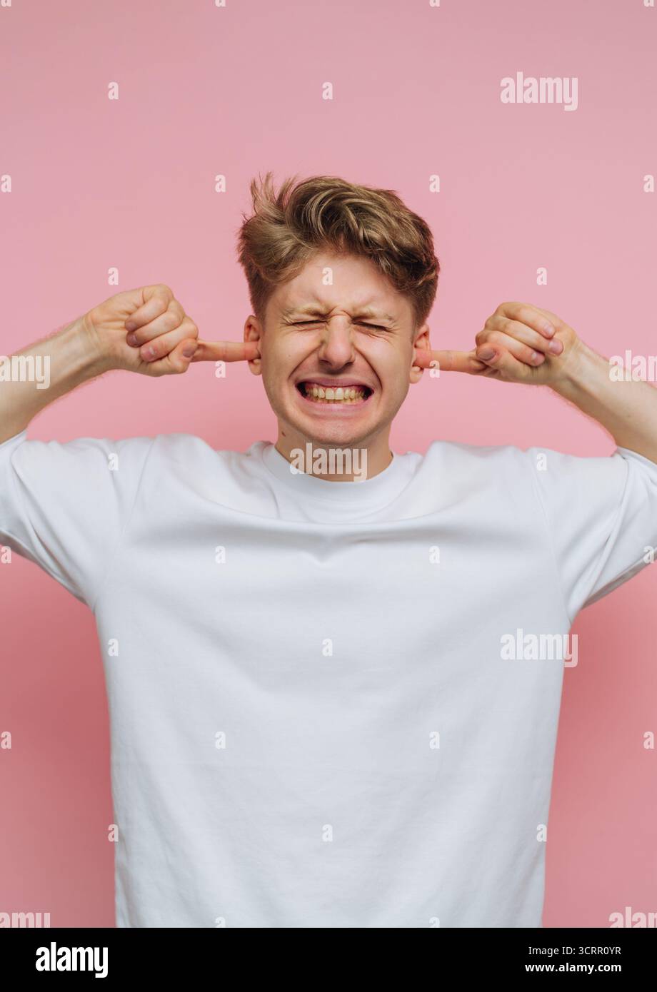 Un jeune homme aux cheveux blonds courts sourit largement en bouchant ses oreilles avec ses doigts. Il se tient sur un fond rose Uni, véhiculant un humour Banque D'Images
