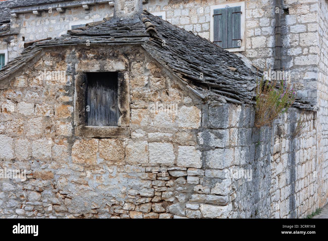 Un bâtiment en pierre altérée avec un toit en ardoise en pente et une petite fenêtre sombre se trouve le long d'un mur de pierre brute. Les plantes sauvages s'accrochent au bord, créant tim Banque D'Images