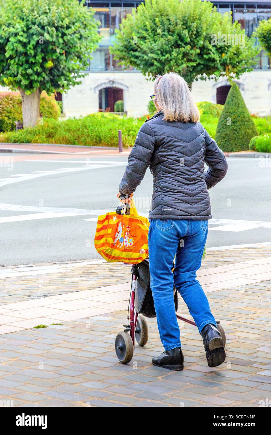 Femme âgée mature en jeans et veste matelassée avec sac à provisions traversant la rue à l'aide d'un cadre de marche à roulettes - Loches, Indre-et-Loire (37), France. Banque D'Images