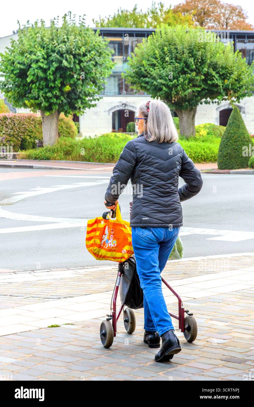 Femme âgée mature en jeans et veste matelassée avec sac à provisions traversant la rue à l'aide d'un cadre de marche à roulettes - Loches, Indre-et-Loire (37), France. Banque D'Images