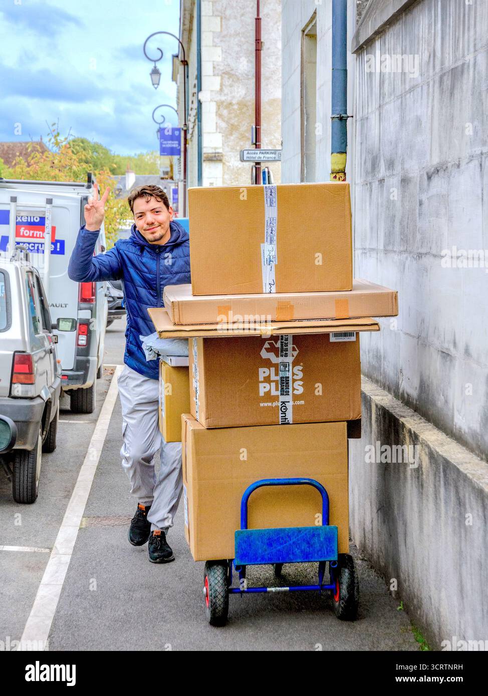 Jeune livreur poussant plusieurs grandes boîtes sur le chariot donnant le signe de victoire lors de la photographie - Loches, Indre-et-Loire (37), France. Banque D'Images