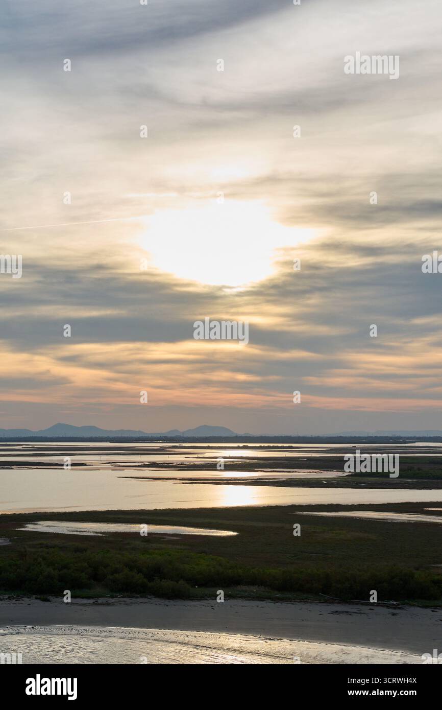 Le soleil se couche sur un vaste paysage humide, créant un reflet lumineux sur les eaux calmes et montrant une silhouette montagneuse lointaine. Banque D'Images