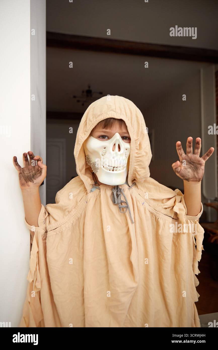 Enfant dans le cape d'Halloween effrayant et le masque de mâchoire de crâne montrant le geste effrayant tout en regardant l'appareil photo à la maison et en s'amusant pendant les fêtes celebbratio Banque D'Images Enfant dans le cape d'Halloween effrayant et le masque de mâchoire de crâne montrant le geste effrayant tout en regardant l'appareil photo à la maison et en s'amusant pendant les fêtes celebbratio Banque D'Images