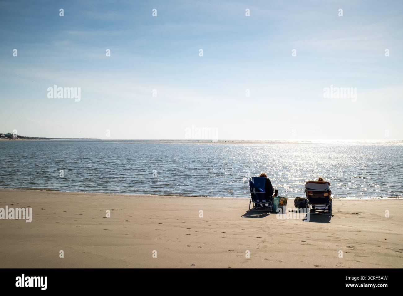 Chaises de plage sur Isle of Palms Beach près de Sullivans Island Caroline du Sud // SULLIVANS ISLAND, Caroline du Sud — deux personnes se détendent dans des chaises de plage sur la rive sablonneuse de Sullivans Island, une île barrière dans le comté de Charleston. Les eaux calmes de l'océan Atlantique reflètent l'éclat du soleil, s'étendant vers l'horizon. Au loin, le littoral de l'île de Palms est visible à travers Breach Inlet. Sullivans Island est connue pour son phare historique et sa proximité avec Charleston, une grande ville portuaire. Banque D'Images