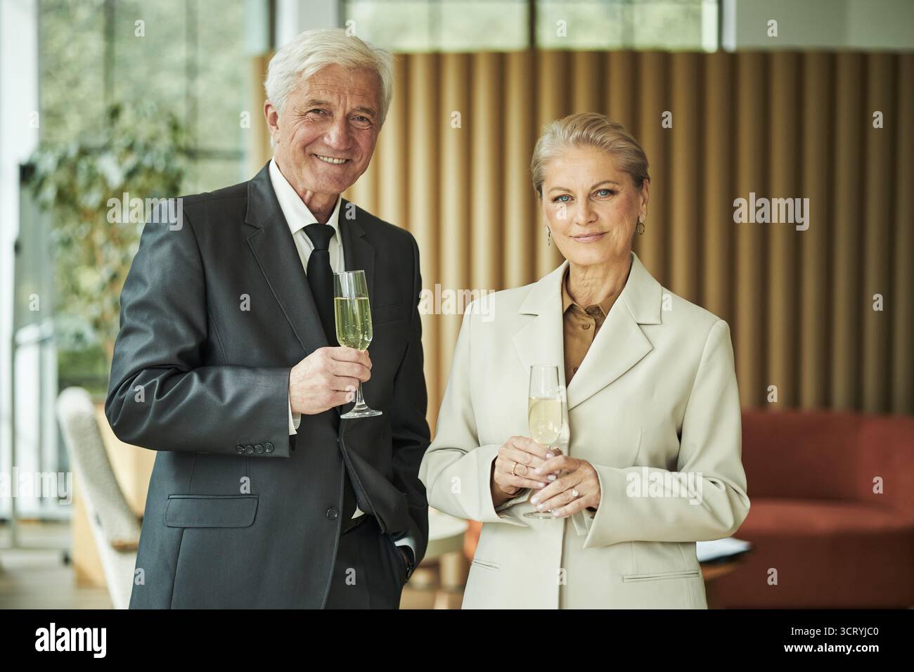 Homme caucasien senior et femme caucasienne senior debout ensemble tenant des verres de champagne, souriant à la caméra dans un cadre intérieur moderne, célébrant une occasion spéciale Banque D'Images