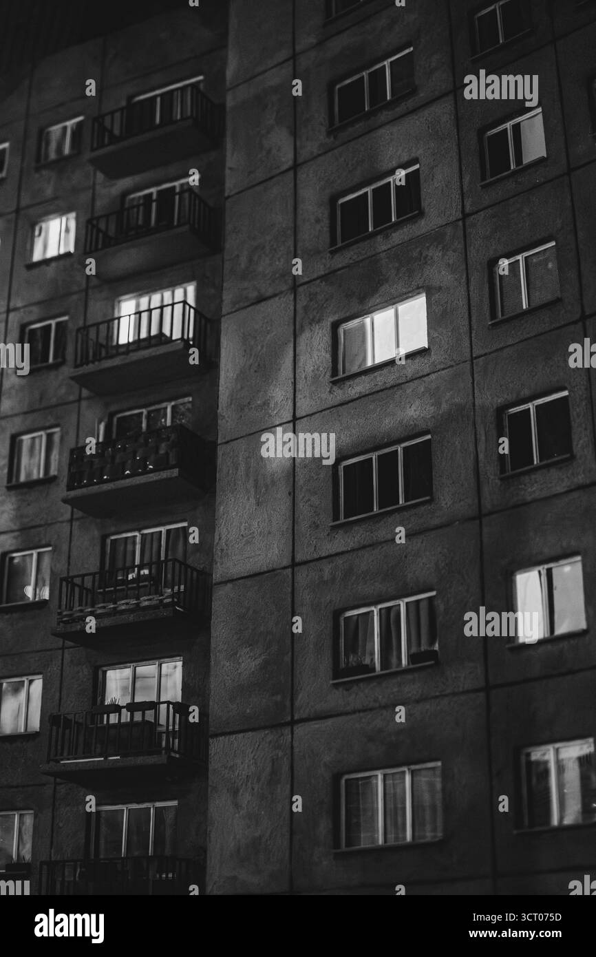 Photo en noir et blanc d'un immeuble nocturne de l'ère socialiste avec des fenêtres éclairées, évoquant la solitude et l'aliénation urbaine. Banque D'Images