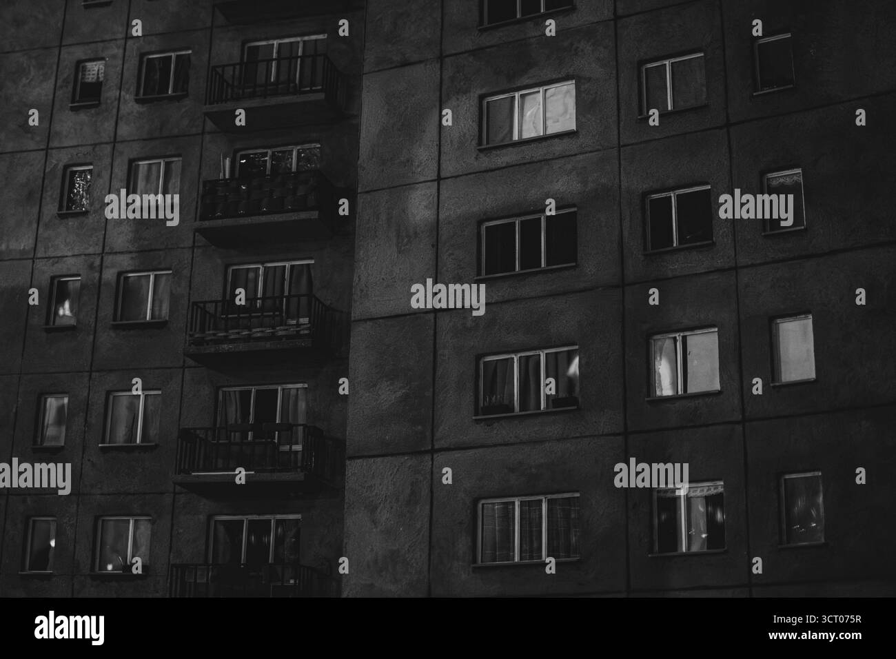 Photo en noir et blanc d'un immeuble nocturne de l'ère socialiste avec des fenêtres éclairées, évoquant la solitude et l'aliénation urbaine. Banque D'Images