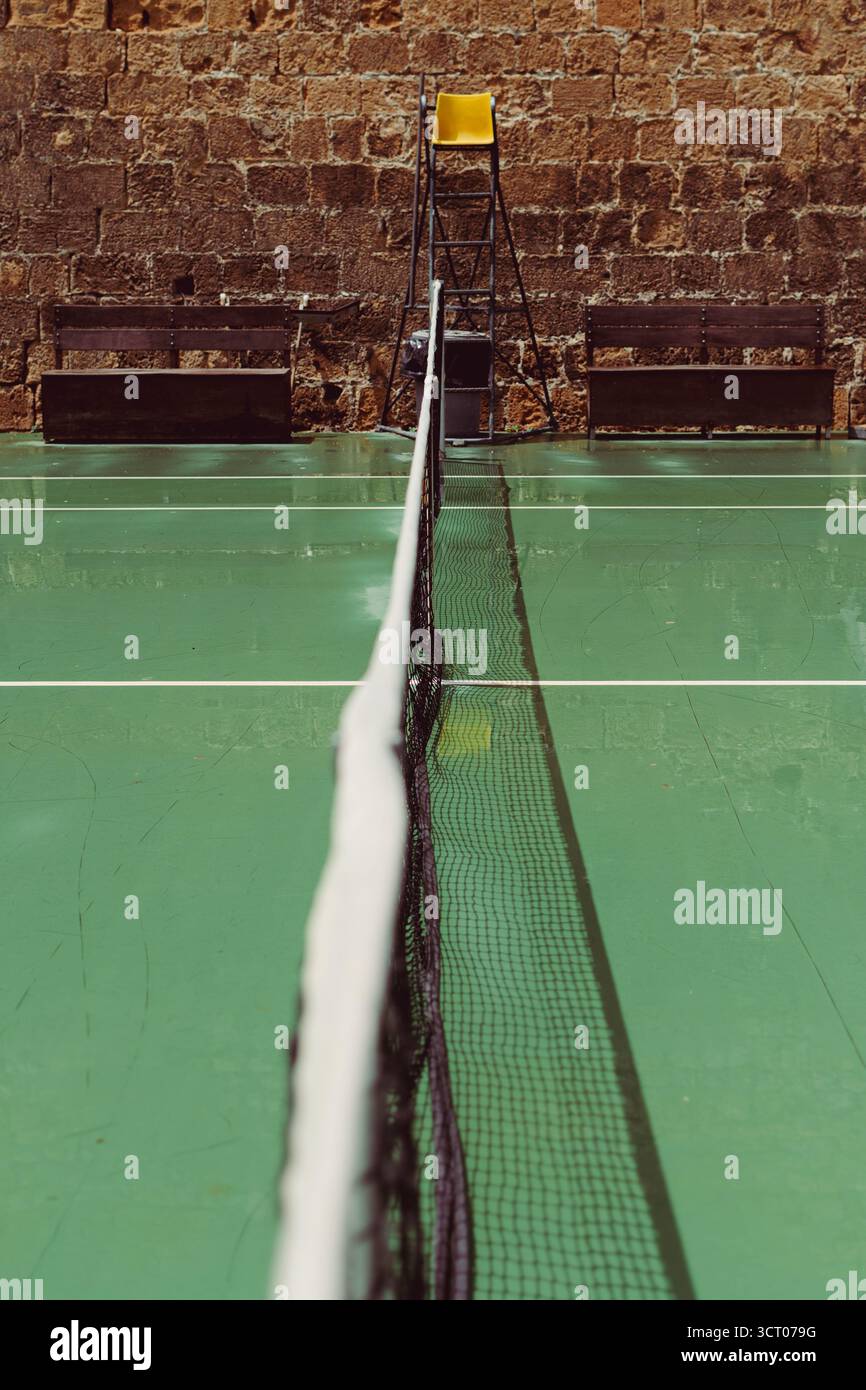 Court de tennis à Héraklion, en Grèce, avec un filet centré, un fauteuil arbitre jaune et un mur de pierre en toile de fond, capturant un mélange tranquille de sport et d'archite âgé Banque D'Images