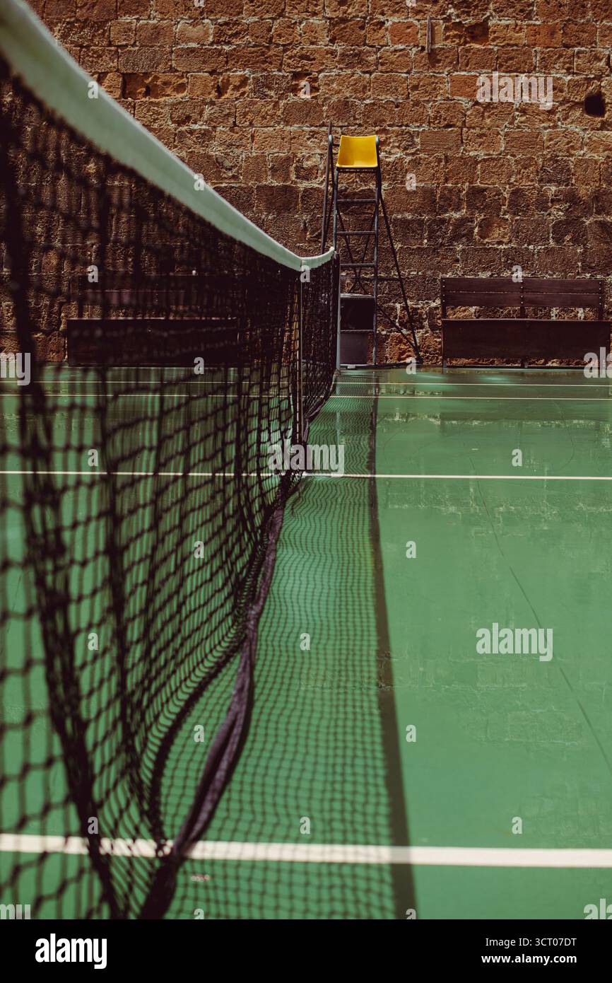 Court de tennis à Héraklion, en Grèce, avec un filet centré, un fauteuil arbitre jaune et un mur de pierre en toile de fond, capturant un mélange tranquille de sport et d'archite âgé Banque D'Images