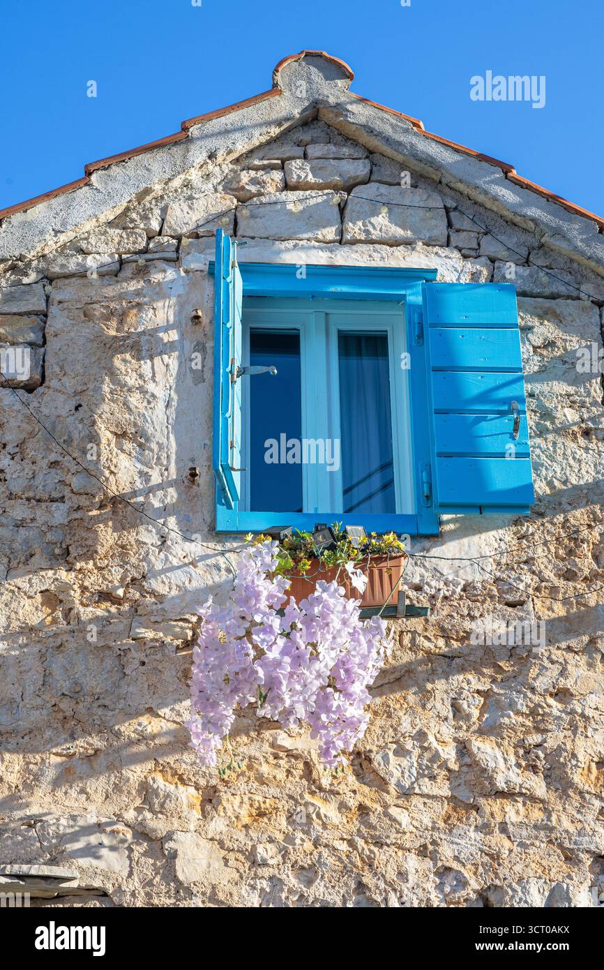 Charmant cottage en pierre ensoleillé avec volets bleus vifs, une fenêtre de fleurs blanc-violet, des murs rustiques texturés et un ciel lumineux évoquant Mediter Banque D'Images