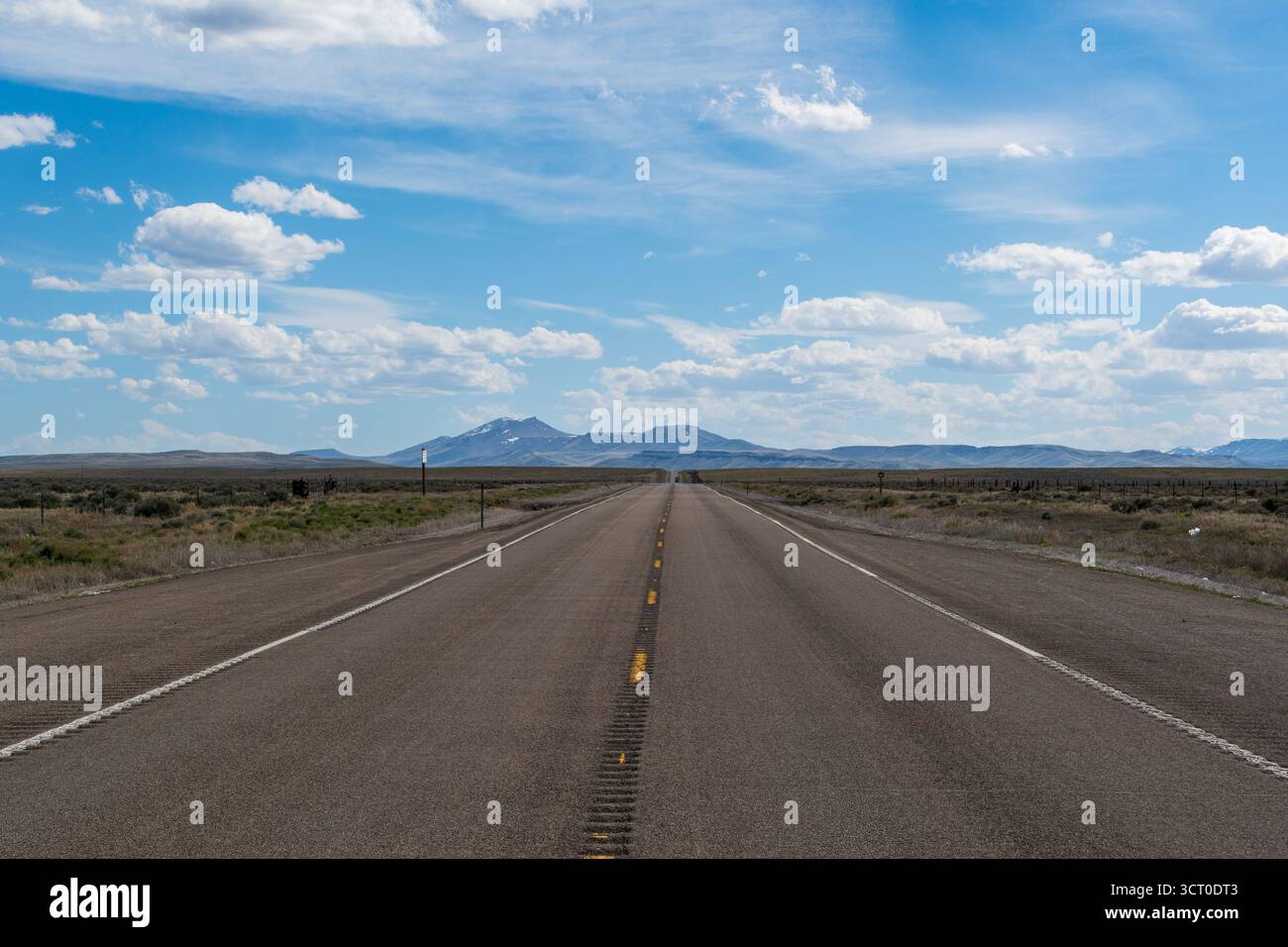 Paysage printanier du Nevada avec route désertique menant vers des montagnes lointaines sous un ciel partiellement nuageux avec des éclats de soleil et des accents bleus. Banque D'Images
