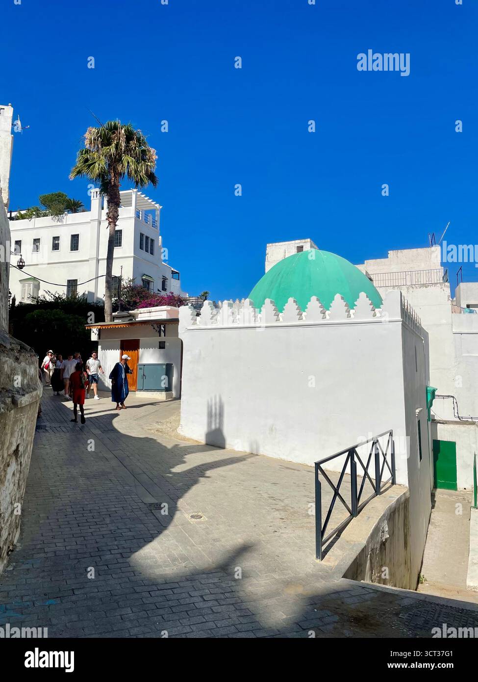 Rue étroite avec des bâtiments blancs et une mosquée verdoyante sous un ciel clair. Tanger, Maroc — 7 juin 2024. Banque D'Images