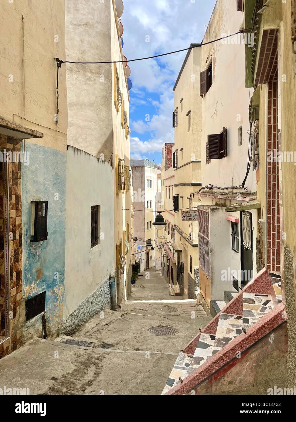 Ruelle en pente dans la vieille médina avec des façades en couches, des marches carrelées et un mélange de textures vieillies sous un ciel partiellement nuageux. Tanger, le 7 juin 2024 Banque D'Images