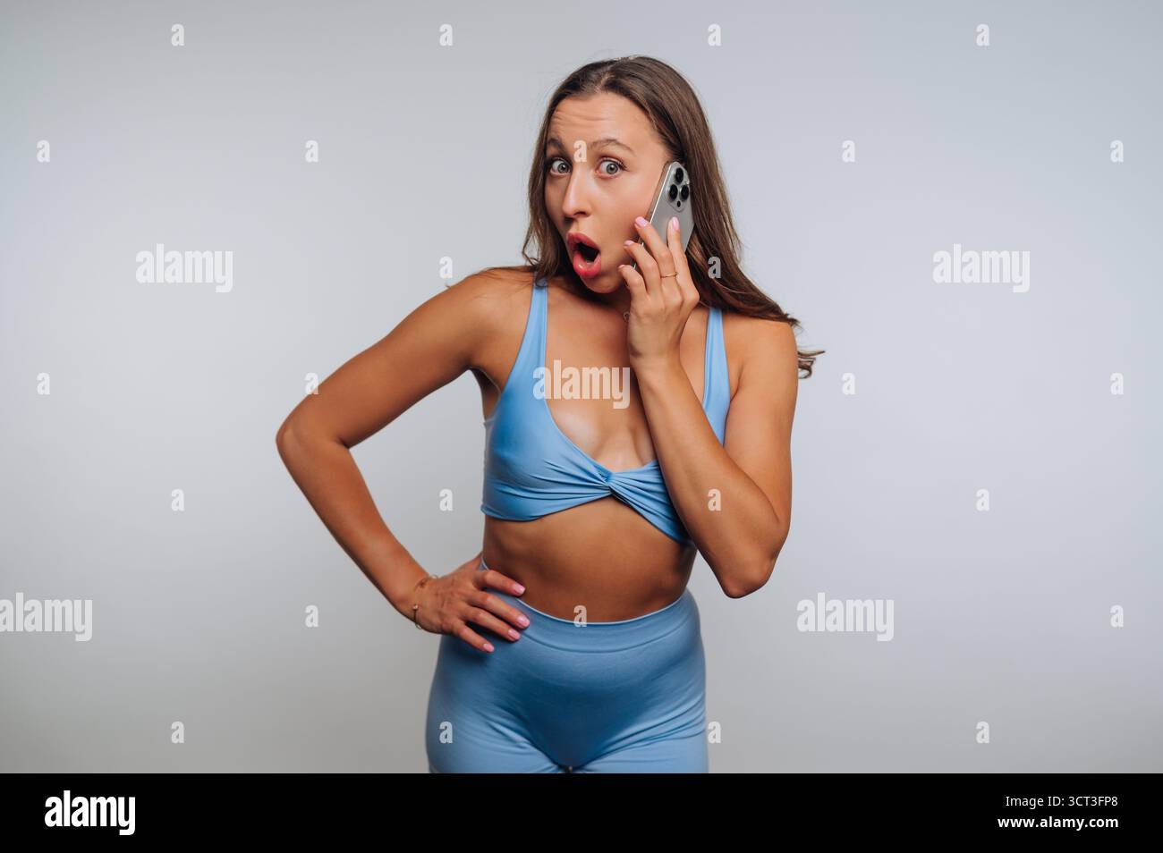 Une femme portant des vêtements de sport bleus se tient debout avec une main sur sa hanche, paraissant surprise en parlant sur son smartphone. Cette action capture son dixième Banque D'Images